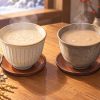 Two cups of warm amazake side by side, one beside a bowl of rice koji and one beside a block of sake lees