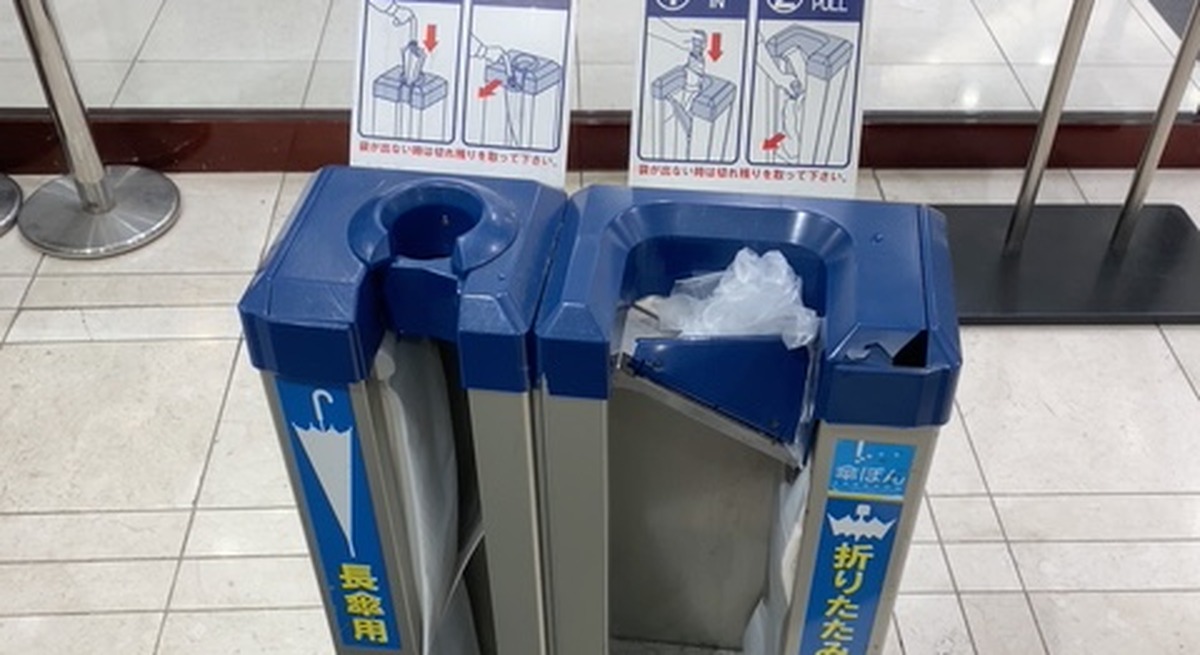 Mechanical umbrella-bagging machine at a building entrance in Japan, with a folded umbrella already wrapped in plastic on the right side.