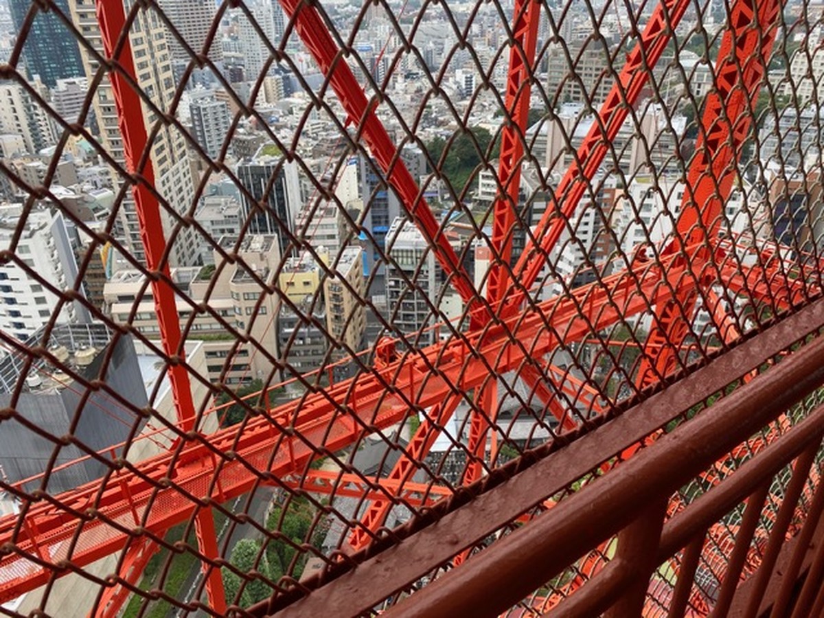 The city below seen through the safety mesh and the tower's orange steel frame, with a stair handrail in the foreground.